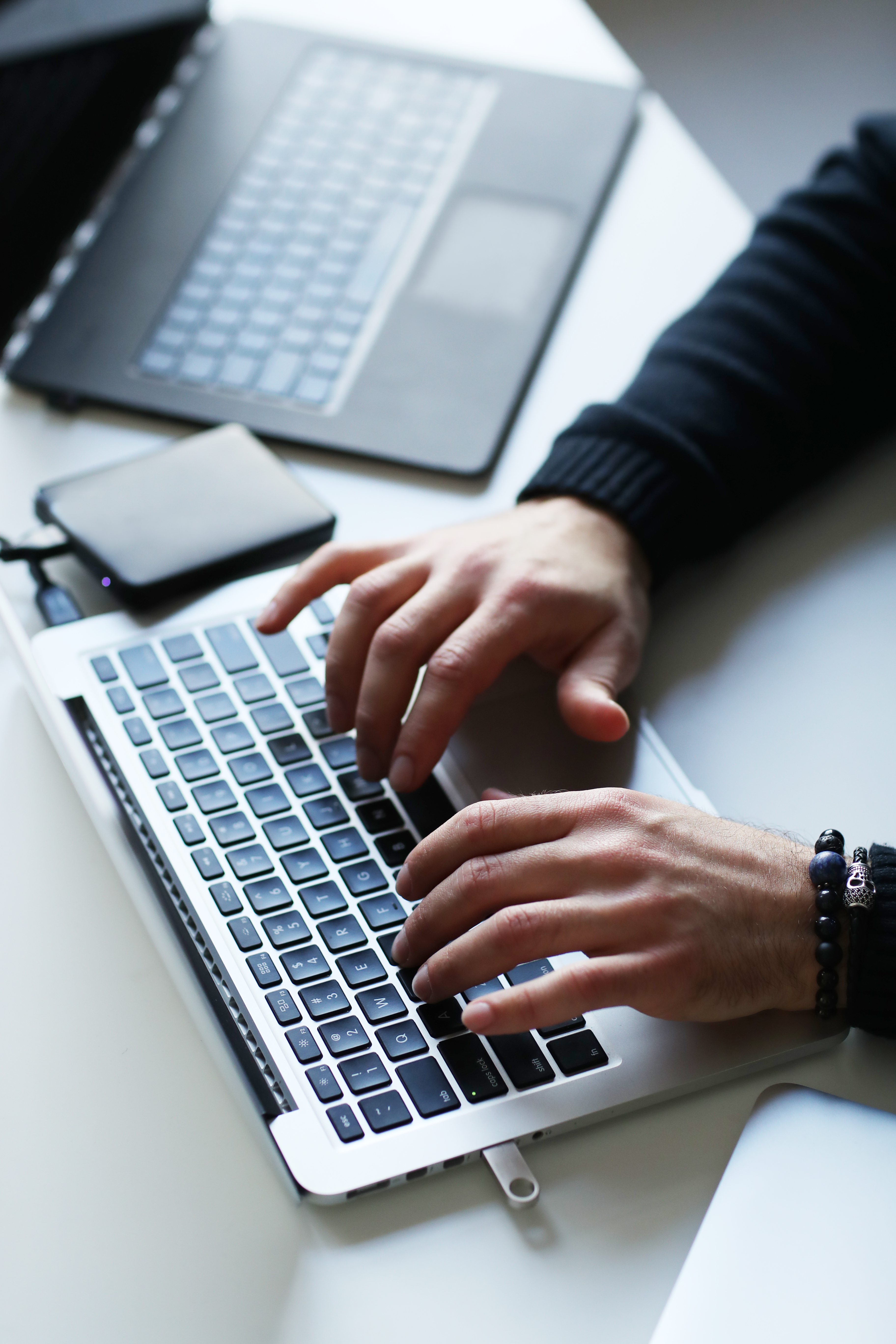 One black laptop is the background and one white laptop in the foreground with a small black hard drive in the middle. A pair of hands are typing on the laptop. A black sleeve and 2 black bracelets with black linked balls are visible.